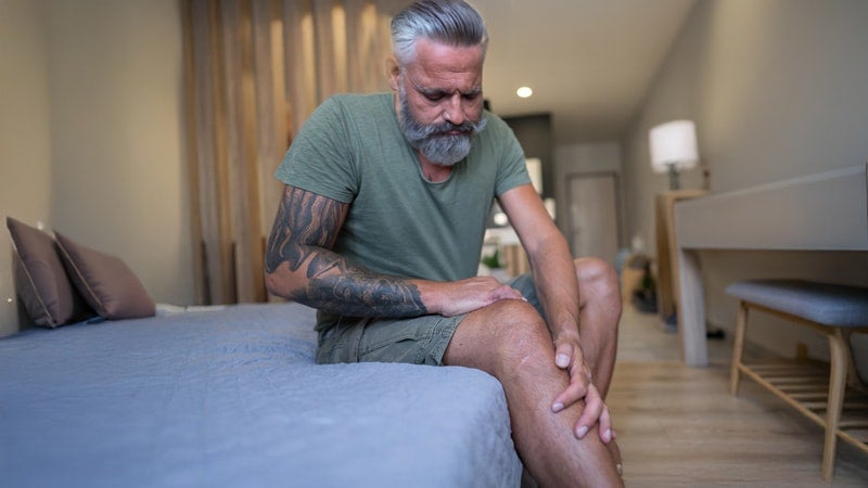 Man on a bed massaging his legs and indicating pain in joints.