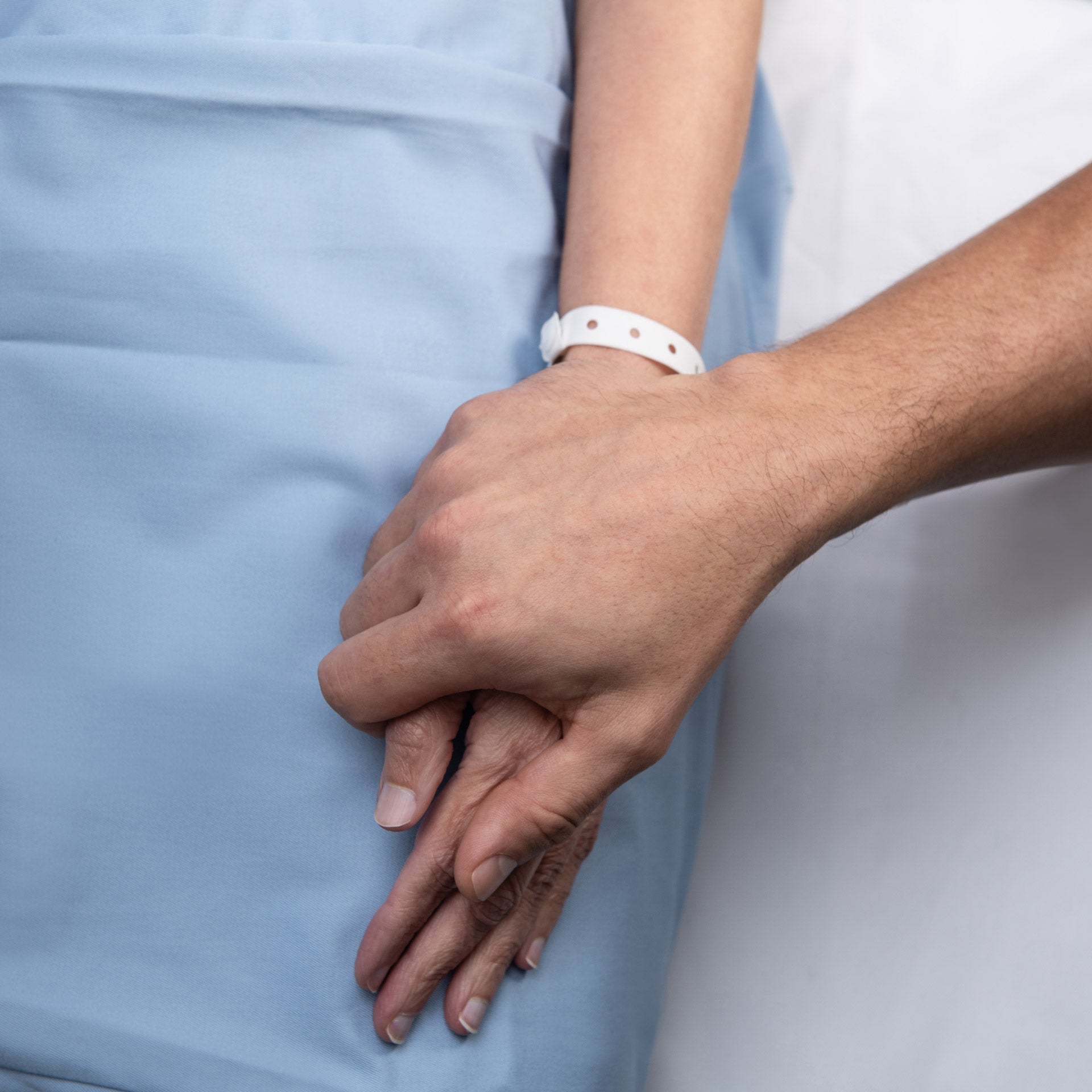 Close-up of a patient lying in a hospital bed wearing a blue gown and white hospital wristband while a caregiver gently holds their hand in a supportive gesture.
