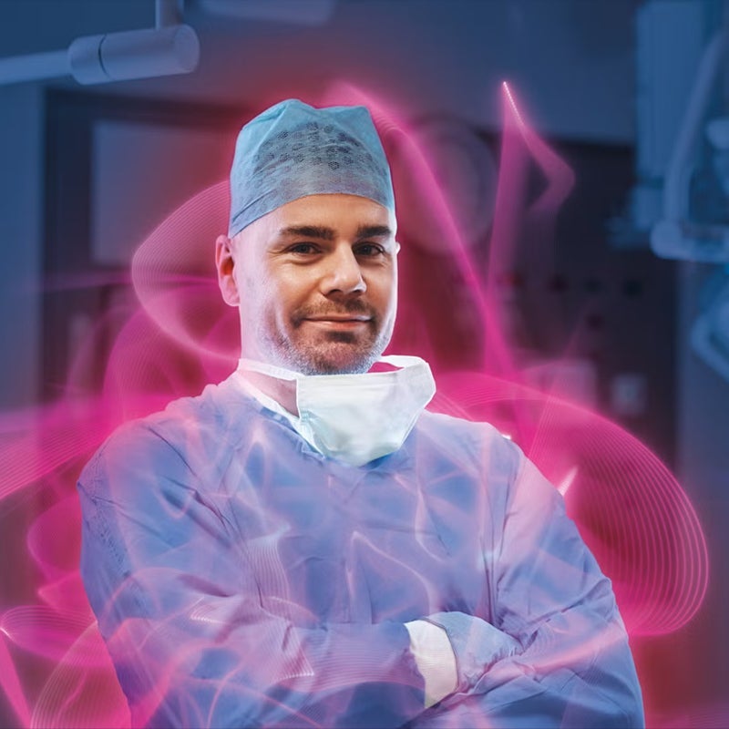 Male surgeon in scrubs with a surgical mask pulled down, smiling with arms crossed, surrounded by a pink glow.