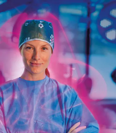 Smiling female surgeon in a surgical cap and scrubs with arms crossed, surrounded by a pink glow in an operating room.
