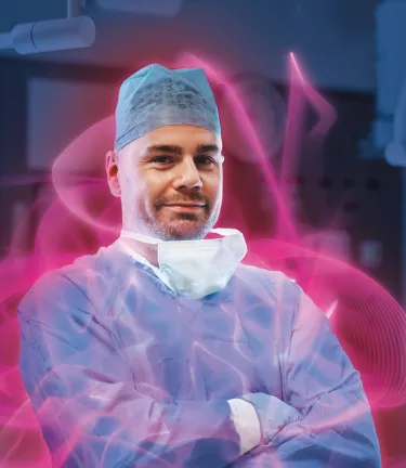 Male surgeon in scrubs with a surgical mask pulled down, smiling with arms crossed, surrounded by a pink glow.