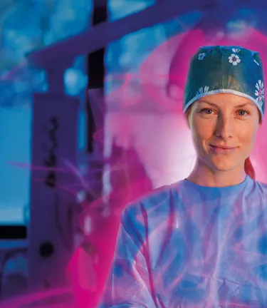 Smiling female surgeon in a surgical cap and scrubs with arms crossed, surrounded by a pink glow in an operating room.